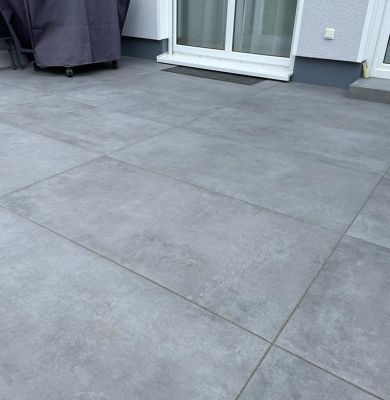 Großformatige graue Terrassenplatten vor einer Tür, links ist ein Grill oder Möbelstück mit Abdeckung zu sehen.