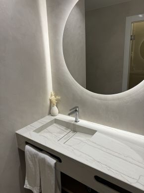 Modernes Bad mit hellem Waschtisch, eingelassenem Waschbecken, rundem Spiegel mit Beleuchtung und Dekovase.